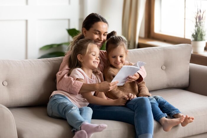 Eine Mutter sitzt mit ihren zwei Kindern auf der Couch uns liest ein Buch vor